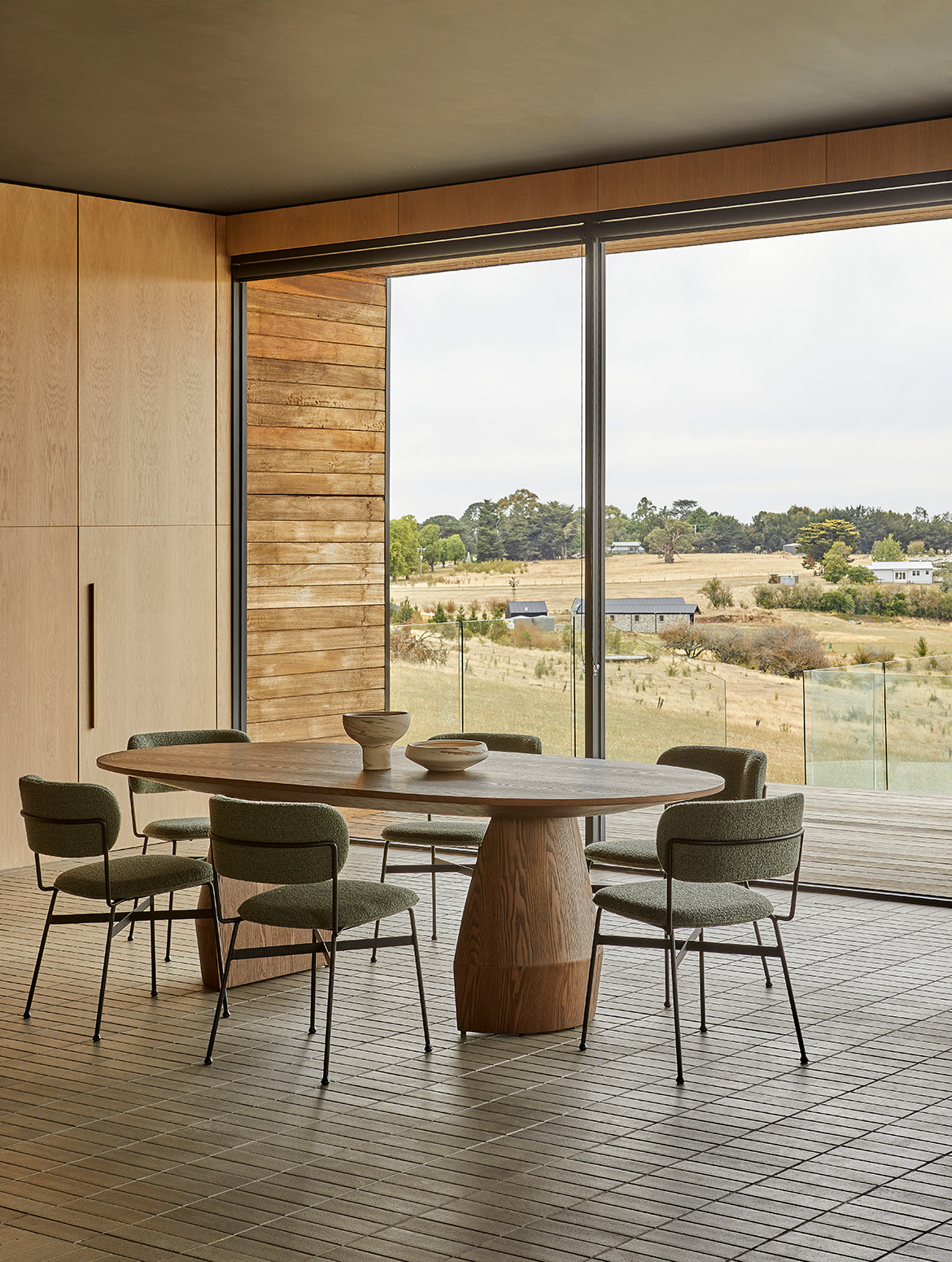 An oval oak dining table on a pedestal base, ringed by green bouclé chairs, looking out over paddocks